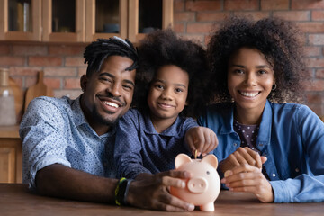Portrait of happy young African American family with teen daughter put coin save money in piggybank. Smiling economical ethnic parents with child feel provident make financial investment for future.