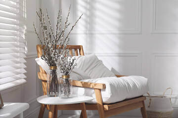 Glass vases with pussy willow tree branches on table near armchair in room, space for text