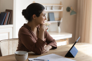 Pensive young mixed race ethnicity woman sit at desk use tablet look in distance dreaming thinking....