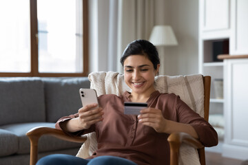 Smiling young Indian woman sit in chair at home shopping online on smartphone with credit debit card. Happy millennial ethnic female buyer make payment buy on internet use secure banking on cellphone. - Powered by Adobe