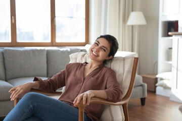 Portrait of smiling young Indian woman sit relax in armchair relieve negative emotions breathe...