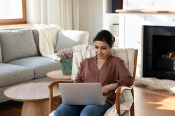 Young Indian woman sit in chair at home browse wireless internet on modern laptop gadget. Mixed race female relax in armchair use computer work or study distant on device. Technology concept.
