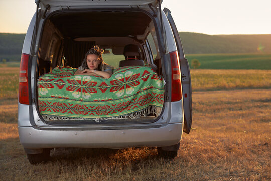 a hipster girl lies in bed in a minivan against the background of the field and the setting sun. The concept of freedom, youth and travel. High quality photo