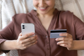 Fototapeta premium Crop close up of happy young Indian woman relax at home make payment purchase on smartphone with credit debit card. Smiling ethnic female buyer customer shop on internet on cellphone from home.
