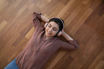Top view of happy young Indian woman wear headphones lying on floor in living room listen to music....