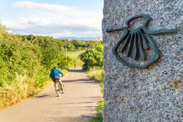 Pilgrim cyclist on the Camino de Santiago (Sant James Way) loaded with a backpack next to a column with the shell symbol of the Camino Santiago de Compostela - obrazy, fototapety, plakaty