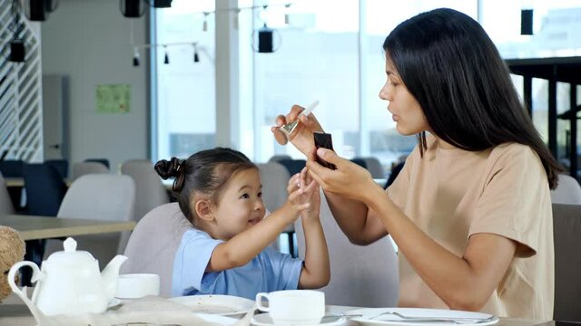 Mother applies make-up on daughter face. Asian woman brunette moves brush with powder to smiling toddler girl cheeks in cafe close view