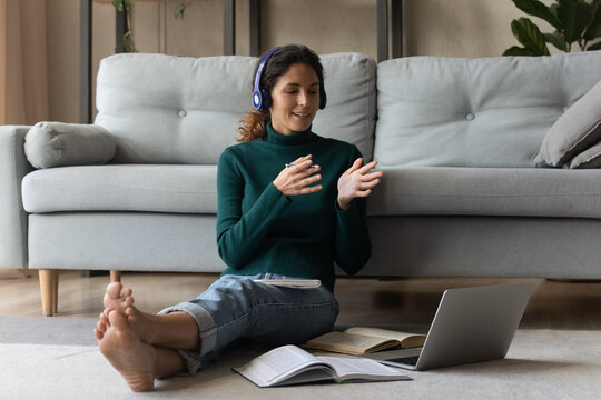 Woman in headphones involved in internet meeting, sitting on warm floor at home, speaking at laptop webcam, teacher leading online lesson, explaining, making video call, student learning language
