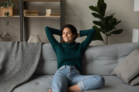 Smiling attractive young woman leaning back with hands behind head on cozy couch, looking to aside, relaxing, enjoying lazy weekend at home, dreaming about good future, visualizing new opportunities