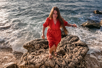 A girl in a red dress on the rocks on the seashore. Sea sunset. Journey.