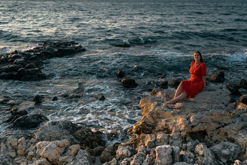 A girl in a red dress on the rocks on the seashore. Sea sunset. Journey.