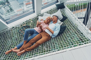Beautiful mature couple examining smart phone and smiling while relaxing in big hammock at home...