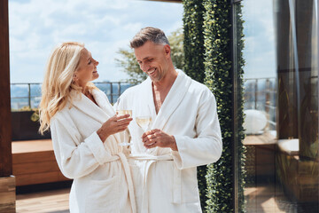 Beautiful mature couple in bathrobes enjoying champagne while relaxing in luxury hotel outdoors - obrazy, fototapety, plakaty