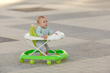 A cute little boy is learning to walk on a walker taking his first step in the park on a bright...