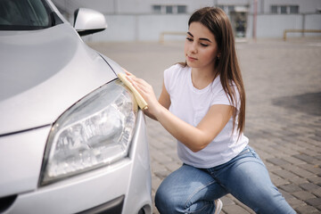 Front view of attractive happy joyful female driver washing her car's headlights with special rag...