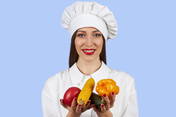 A young girl cook in a white robe and a cap stands and holds a vegetables isolated on a blue background.