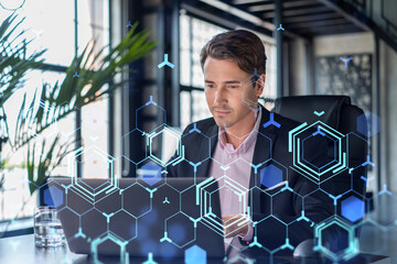 Handsome businessman in suit at workplace working with laptop to optimize development by implying new technologies in business process. Hi tech hologram over office background