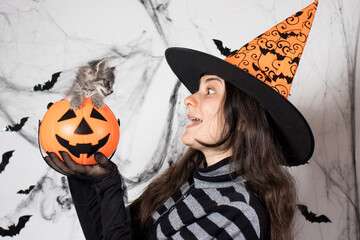 A woman in a witch costume in a hat holds a bucket pumpkin with cat. Halloween people and animal.