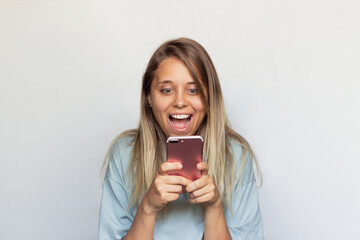 A young caucasian excited, astonished and delighted blonde woman smiles, holding a mobile phone and looking at the screen isolated on a light color gray background. The girl uses her smartphone