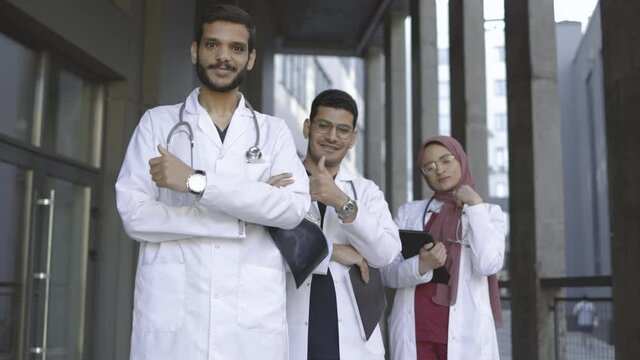 Portrait of three young Arabic Malay medical students team showing thumbs up while standing in a row outside modern clinic or university campus