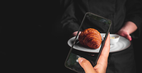 woman hand holding and showing smart phone takes a photo chef in a black suit holds in his hands plate with croissant on Dark grey black background. food photos