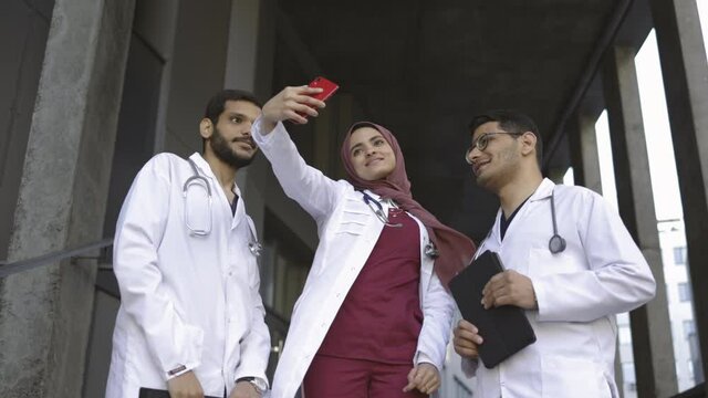 Group of three young doctors interns or medical students taking selfies while standing outdoors of the hospital. Arabian medical team making selfie photo or video call on smartphone outside