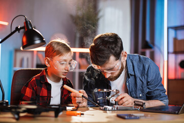 Caucasian father and his son in protective glasses using soldering iron for fixing display card from laptop. Two men sitting together at table and repairing modern technology at home.