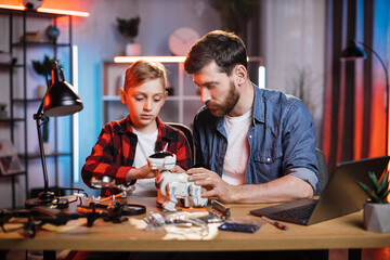 Caring father controlling repairing process of toy robot dog by his little son. Bearded man teaching boy fixing broken appliances at home. Educational moments during parenthood.
