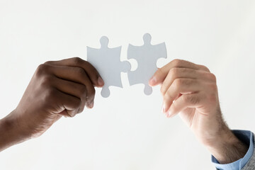 Crop close up of two diverse multiracial people hold puzzle pieces look for business solution together. Multiethnic employees connect jigsaw parts solve problem engaged in teambuilding activity.