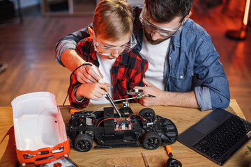Father with son in protective glasses using soldering iron for repairing red toy car at home....