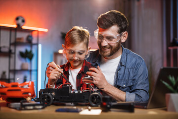 Happy young father teaching his little son for soldering remote controlled car at home. Man and boy wearing protective glasses while fixing broken toy.