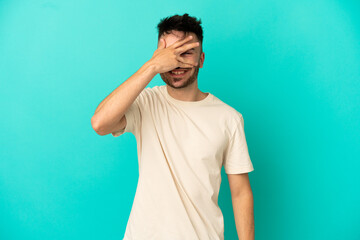 Young caucasian man isolated on blue background covering eyes by hands and smiling