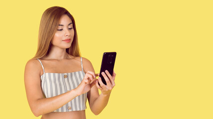 Portrait of beautiful latina girl. She uses cell phone happily on yellow background.