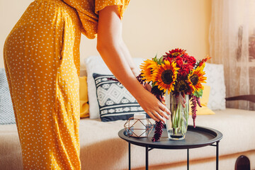 Woman puts vase with sunflowers and zinnia flowers on table. Housewife takes care of interior and fall decor at home. - Powered by Adobe