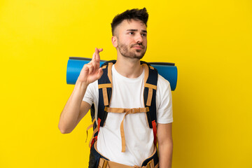 Young mountaineer caucasian man with a big backpack isolated on yellow background with fingers crossing and wishing the best