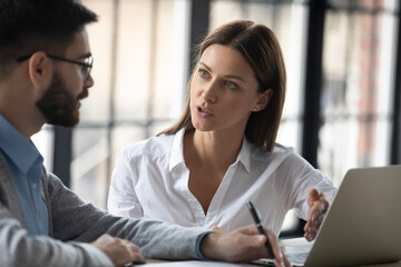Serious diverse businesspeople sit at desk cooperate brainstorm on computer together at team meeting. Focused man and woman colleagues talk speak discuss company business ideas work on laptop.