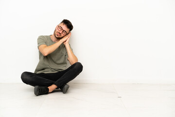 Young caucasian man sitting on the floor isolated on white background making sleep gesture in dorable expression