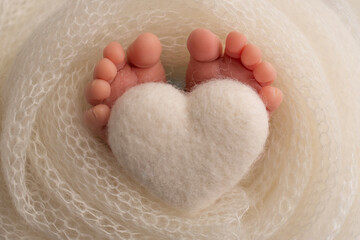 The feet of a newborn baby are wrapped in a knitted blanket. The fingers of a newborn baby are holding a white knitted heart.