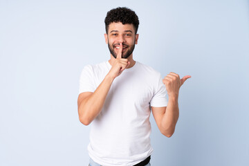 Young Moroccan man isolated on blue background pointing to the side and doing silence gesture