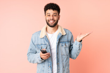 Young Moroccan man using mobile phone isolated on pink background with shocked facial expression