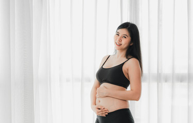 Smiling two month pregnant Asian woman touch her belly by the window