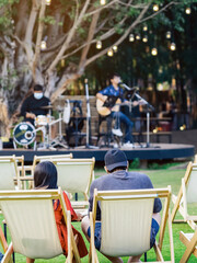 Back view of Asian young couple sit to relax on white deck chairs with tables for dinner in lawn is surrounded by shady green grass with blurred image of musical performance on stage in background.