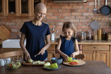 Happy daughter child helping cancer patient mom to cook dinner. Sick hairless mother and girl preparing healthy organic salad from fresh natural ingredients, cutting vegetables on kitchen table
