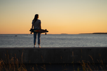 Young fashion girl with longboard enjoy sunset looking at seaside skyline. Stylish trendy female longboarder silhouette over beautiful setting sun at sea coast. Freedom, youth and lifestyle concept