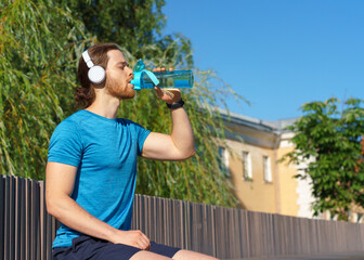 Thirsty young sportsman in blue shirt drinking fresh water from bottle while standing outdoors with green trees on background, active man in headphones taking break. Water balance during workout