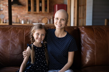 Happy mom with hairless head hugging cute little daughter girl at home, enjoying leisure time with kid on sofa, smiling at camera. Woman going through cancer with support of family, Headshot portrait