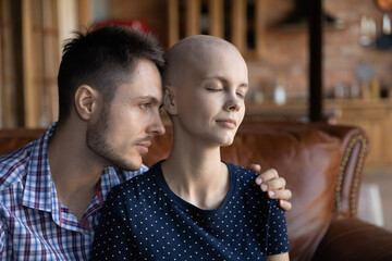 Frustrated exhausted young couple fighting against woman cancer, sitting on couch, hugging, looking away with hope. Husband hugging tired hairless wife with oncology disease, giving support