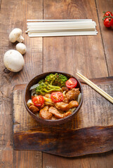 Udon with chicken in Japanese sauce on a plate of coconut shells on a stand next to vegetables and noodles.
