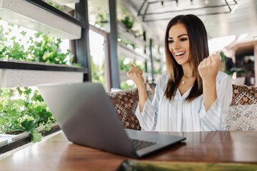 Young woman rejoices in winning having raised her hands up sitting in front of laptop in cafe