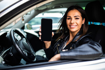 A young beautiful woman driving a car, she points to blank phone screen and smiles. Mobile apps for driver.